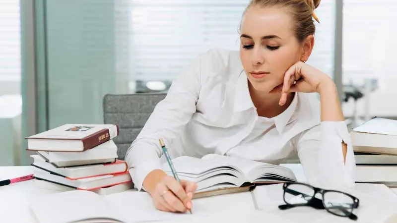 Joven escribiendo en un cuaderno entre libros apilados y unas gafas sobre la mesa