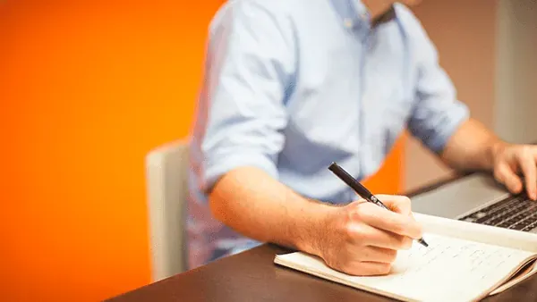 Hombre con camisa azul escribiendo a mano en una libreta frente a un ordenador portátil, sobre un fondo naranja